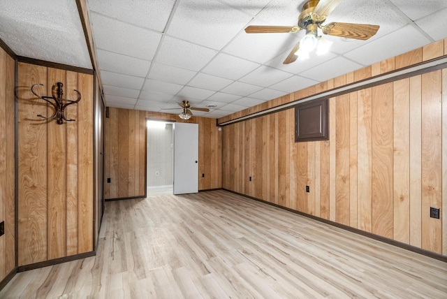 below grade area with ceiling fan, a paneled ceiling, wooden walls, baseboards, and light wood-style floors