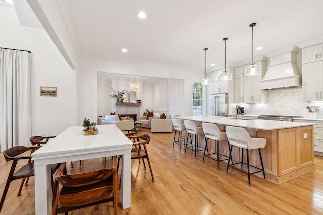 kitchen featuring a warm lit fireplace, a kitchen island with sink, premium range hood, stainless steel built in fridge, and decorative backsplash