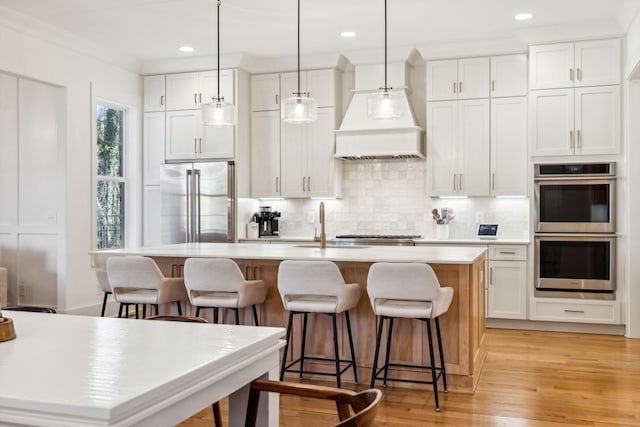 kitchen featuring light countertops, appliances with stainless steel finishes, decorative backsplash, an island with sink, and custom range hood