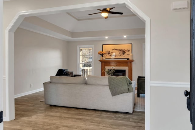 living room featuring a tray ceiling, a ceiling fan, a glass covered fireplace, wood finished floors, and baseboards