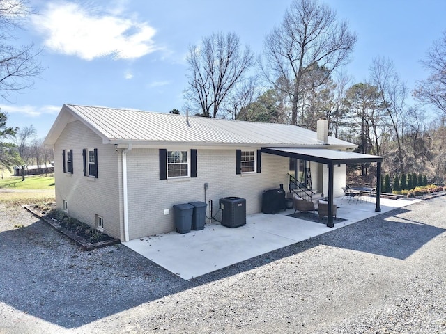 rear view of house with metal roof, a patio, central air condition unit, brick siding, and a chimney