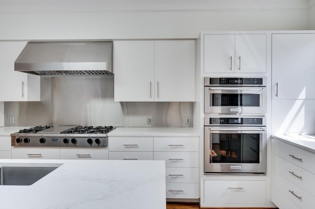 kitchen featuring appliances with stainless steel finishes, white cabinetry, wall chimney range hood, and light stone counters