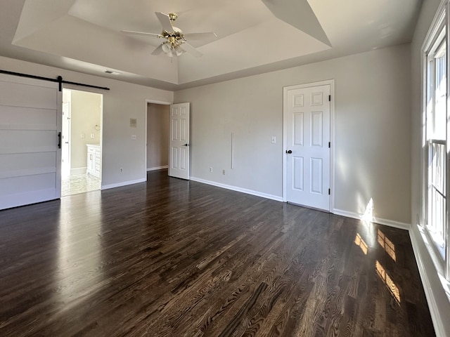 unfurnished bedroom featuring ceiling fan, a barn door, baseboards, dark wood-style floors, and a raised ceiling