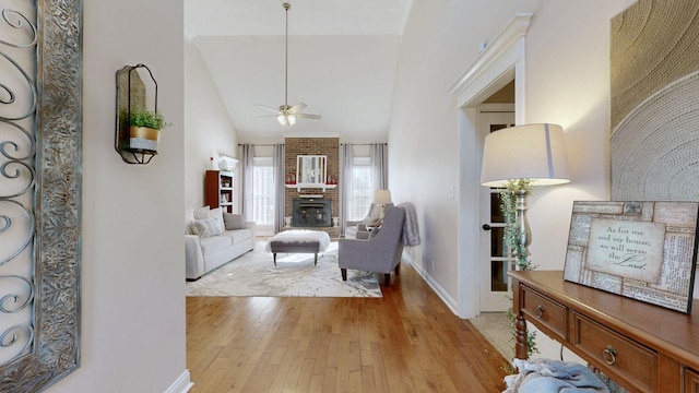 living room featuring a brick fireplace, baseboards, ceiling fan, light wood-style floors, and high vaulted ceiling