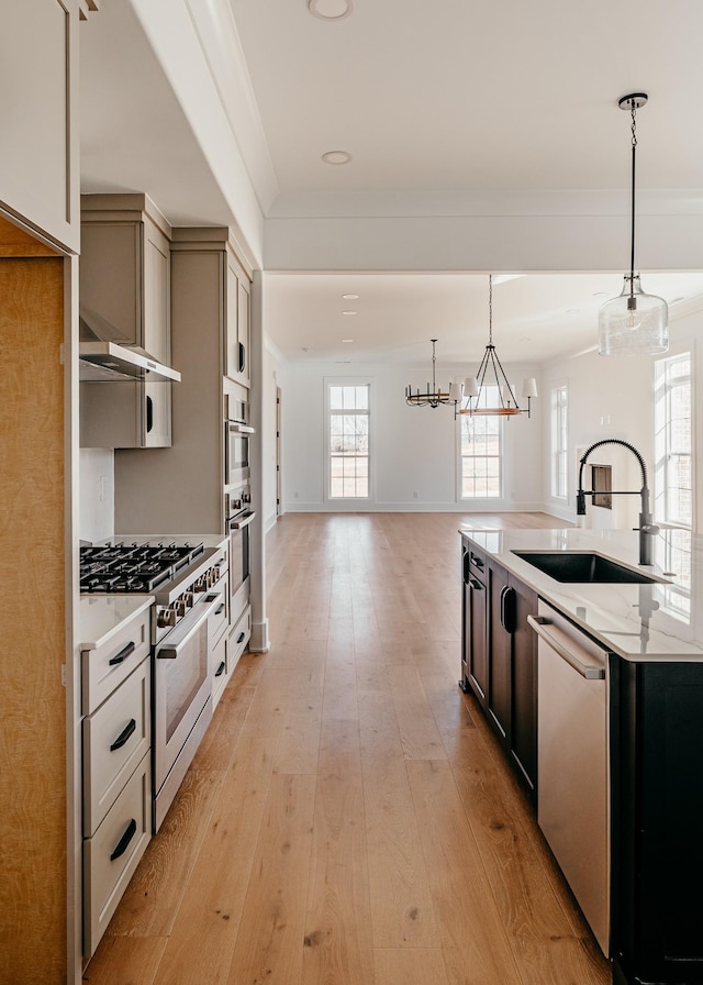 kitchen with a sink, hanging light fixtures, appliances with stainless steel finishes, wall chimney exhaust hood, and light wood finished floors