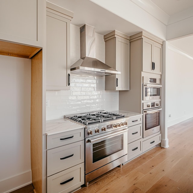 kitchen with backsplash, appliances with stainless steel finishes, light wood-style floors, light stone countertops, and wall chimney exhaust hood