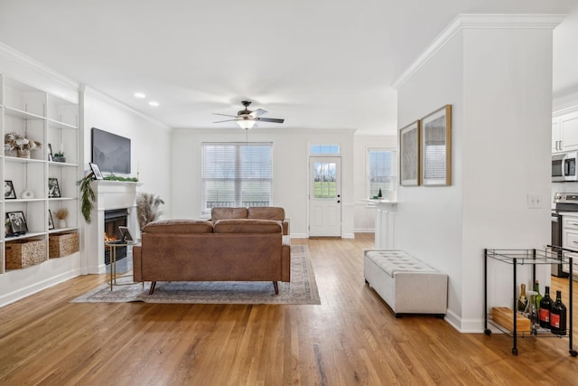 living room with light wood finished floors, ornamental molding, a ceiling fan, a warm lit fireplace, and baseboards