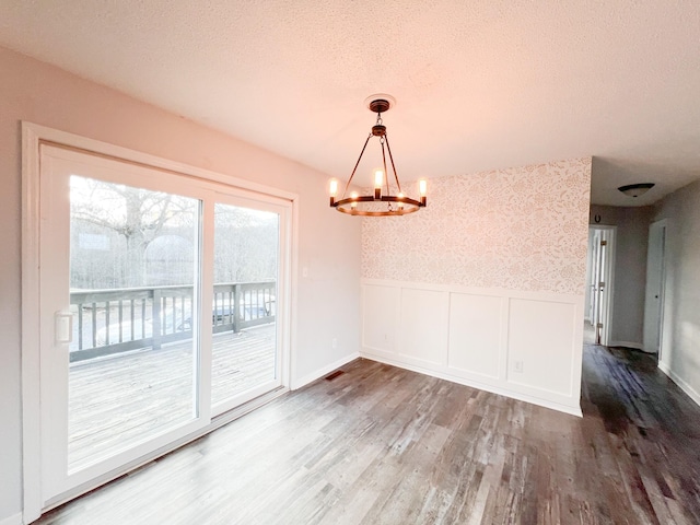 unfurnished dining area featuring an inviting chandelier, wainscoting, a textured ceiling, wood finished floors, and wallpapered walls