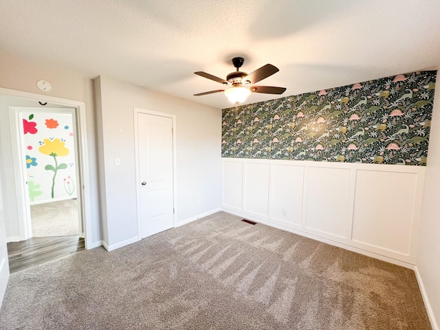 carpeted empty room featuring a textured ceiling, ceiling fan, a wainscoted wall, visible vents, and wallpapered walls