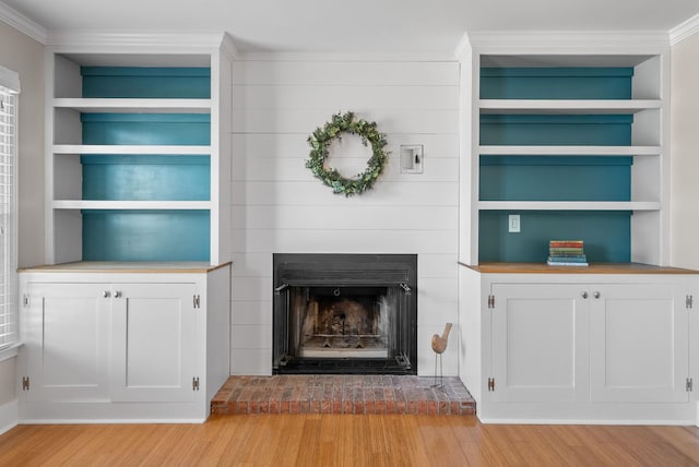 unfurnished living room with built in features, a fireplace with raised hearth, crown molding, and light wood-style flooring