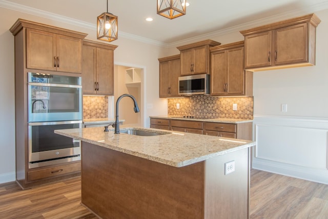 kitchen featuring appliances with stainless steel finishes, light stone counters, ornamental molding, a kitchen island with sink, and light wood-style floors