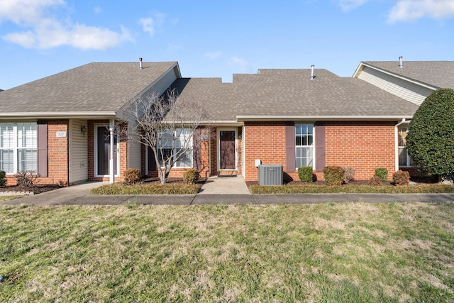 ranch-style home featuring a front yard, brick siding, roof with shingles, and central AC unit