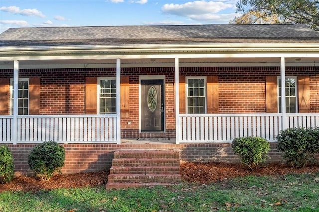 entrance to property featuring covered porch, a shingled roof, and brick siding