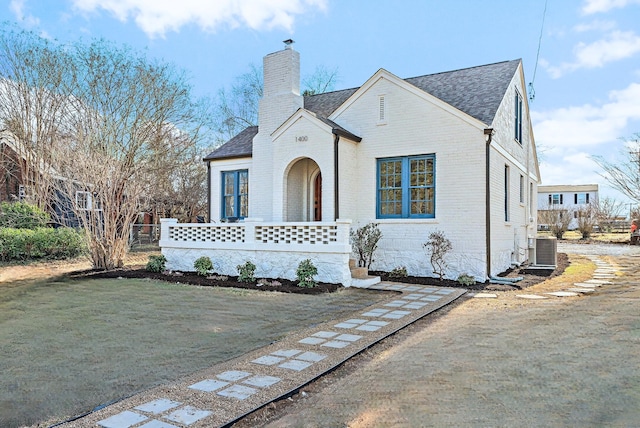 tudor house featuring central air condition unit, brick siding, roof with shingles, a front lawn, and a chimney