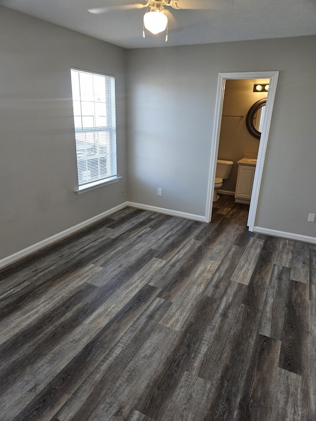 unfurnished bedroom featuring ensuite bath, baseboards, dark wood finished floors, and a ceiling fan