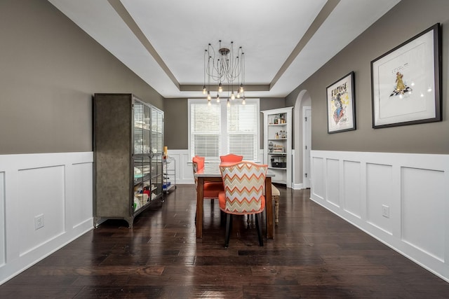 dining space featuring arched walkways, a decorative wall, a tray ceiling, dark wood finished floors, and an inviting chandelier