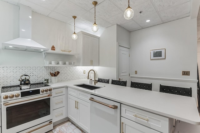 kitchen featuring light countertops, a sink, wall chimney range hood, white appliances, and a peninsula