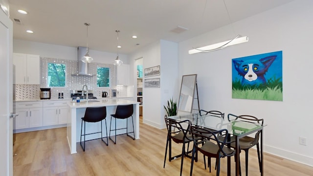 kitchen featuring a kitchen island with sink, white cabinetry, light countertops, backsplash, and wall chimney exhaust hood