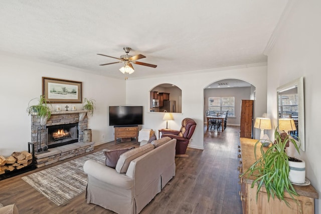 living room featuring dark wood-style floors, crown molding, a ceiling fan, a stone fireplace, and baseboards