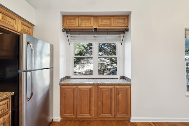 kitchen with wood finished floors, baseboards, brown cabinets, freestanding refrigerator, and light stone countertops