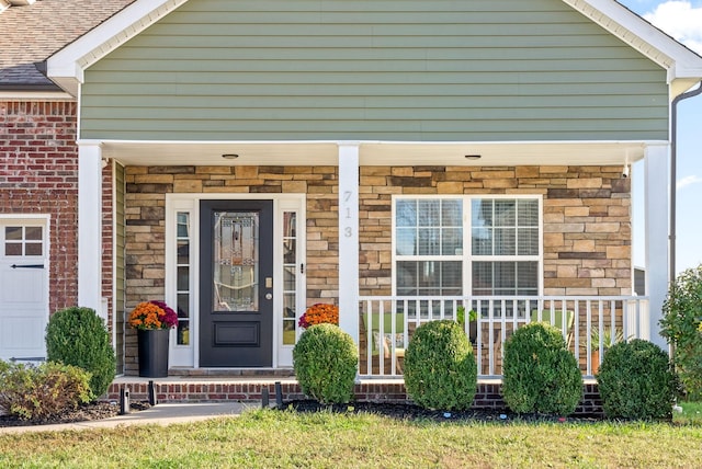 entrance to property with covered porch, stone siding, brick siding, and roof with shingles