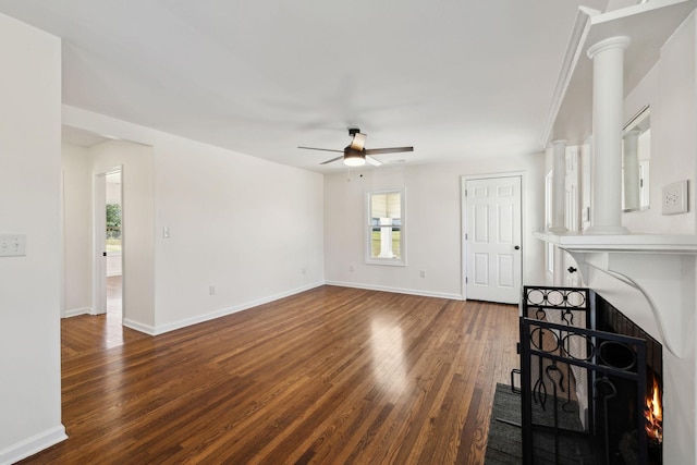 unfurnished living room featuring a warm lit fireplace, ceiling fan, wood finished floors, baseboards, and ornate columns