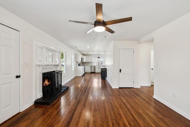 unfurnished living room with a warm lit fireplace, baseboards, a ceiling fan, and dark wood-type flooring