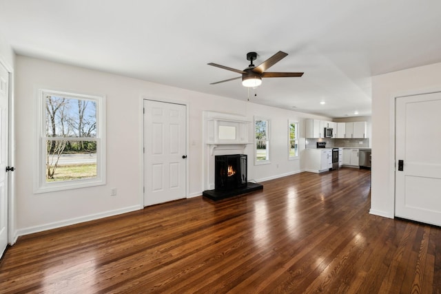 unfurnished living room featuring dark wood-style floors, ceiling fan, a warm lit fireplace, and baseboards