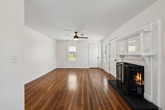 unfurnished living room featuring dark wood-type flooring, a warm lit fireplace, ceiling fan, and baseboards