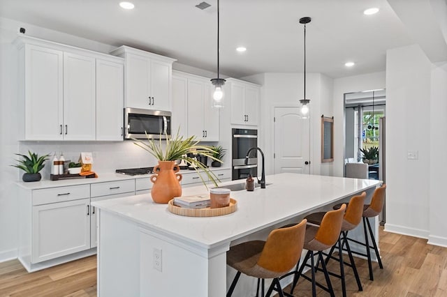 kitchen featuring stainless steel microwave, double wall oven, light wood-style floors, white cabinetry, and a kitchen island with sink