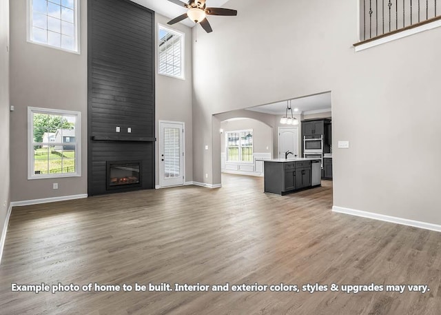 unfurnished living room with arched walkways, a large fireplace, ceiling fan, light wood-type flooring, and baseboards