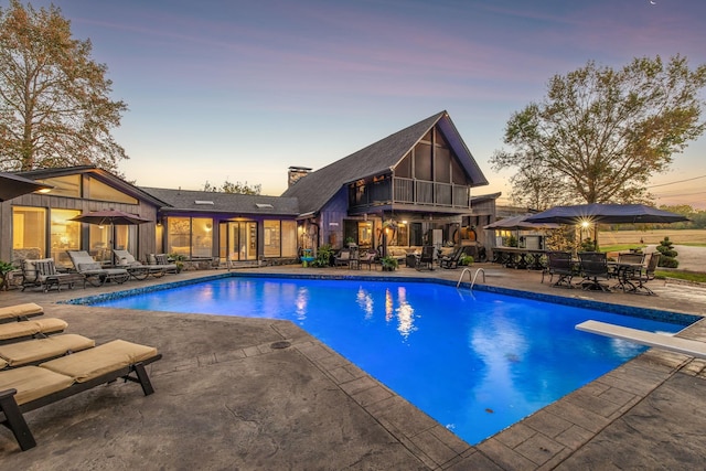 pool at dusk with outdoor dining area, a patio area, an outdoor pool, and a diving board