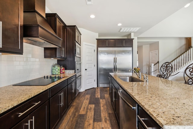 kitchen with visible vents, a sink, light stone countertops, premium range hood, and built in appliances