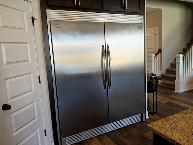 kitchen with dark brown cabinetry, stainless steel built in fridge, dark wood-style floors, and light stone counters