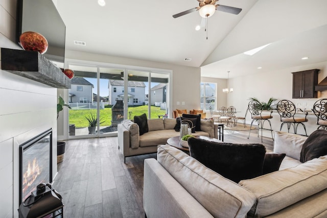 living room with high vaulted ceiling, a tile fireplace, ceiling fan with notable chandelier, wood finished floors, and visible vents