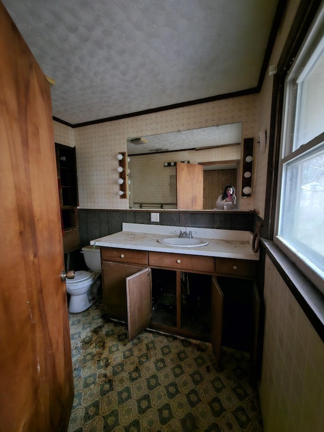 half bathroom with toilet, a wainscoted wall, vanity, ornamental molding, and wallpapered walls