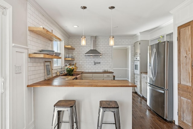 kitchen with ornamental molding, a peninsula, white oven, stainless steel fridge, and wall chimney exhaust hood