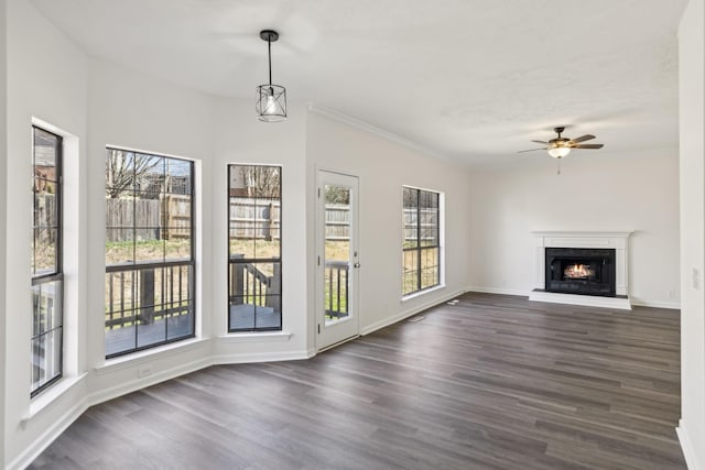 unfurnished living room featuring baseboards, ceiling fan, dark wood-type flooring, a lit fireplace, and crown molding