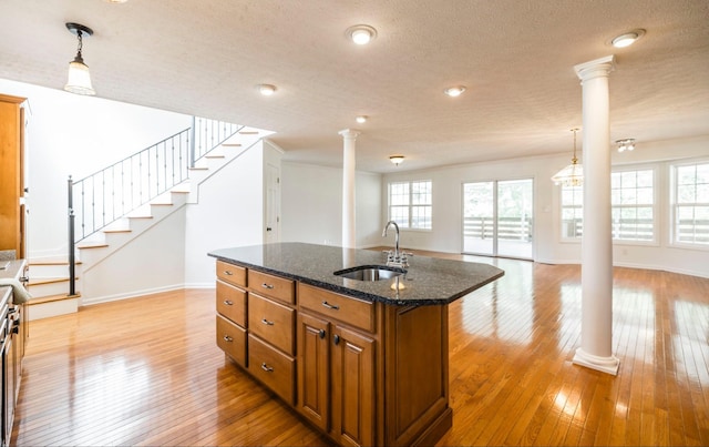 kitchen featuring light wood finished floors, brown cabinetry, a kitchen island with sink, a sink, and ornate columns