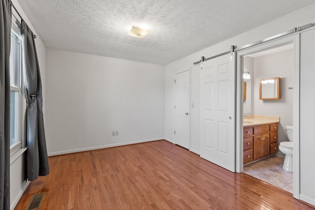 unfurnished bedroom featuring visible vents, a barn door, a textured ceiling, ensuite bath, and light wood-type flooring