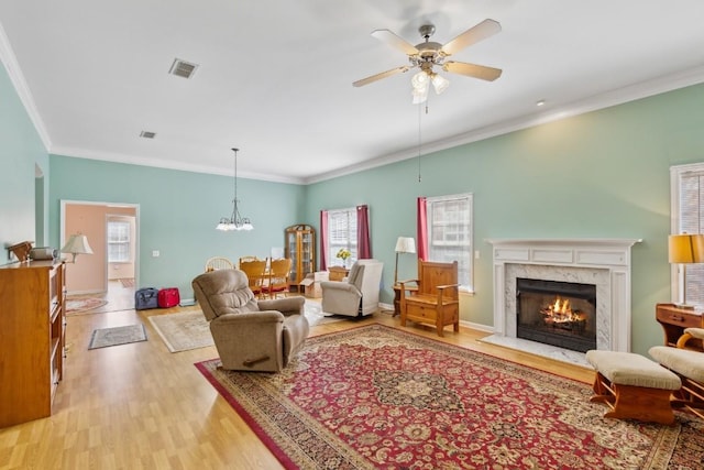 living area featuring a fireplace, visible vents, light wood-style floors, ornamental molding, and ceiling fan with notable chandelier