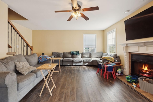 living area with dark wood-style floors, stairway, a warm lit fireplace, and a ceiling fan