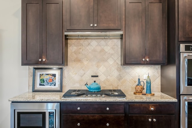 kitchen with appliances with stainless steel finishes, dark brown cabinetry, under cabinet range hood, and light stone countertops