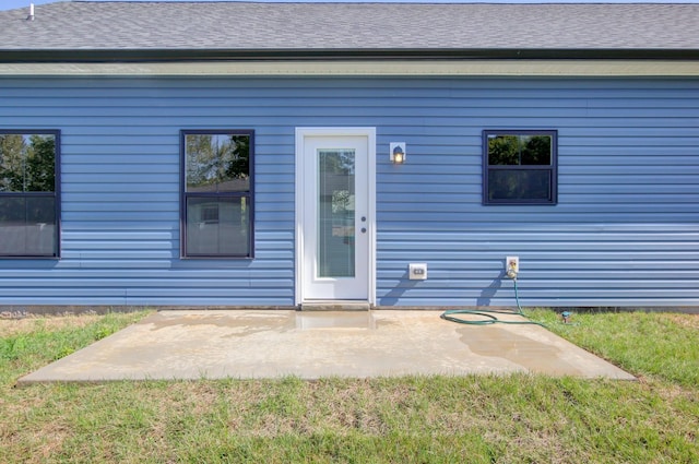 view of exterior entry with roof with shingles and a patio