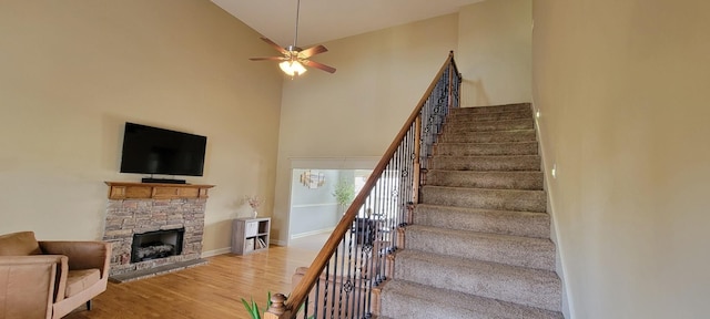 stairs with ceiling fan, a stone fireplace, wood finished floors, a towering ceiling, and baseboards