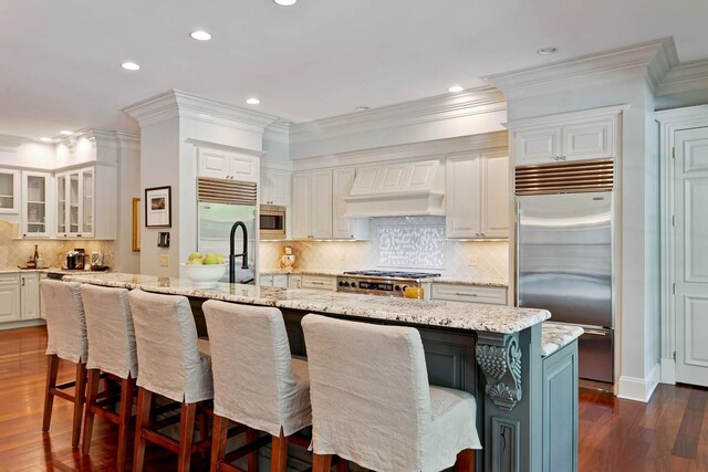 kitchen featuring built in appliances, white cabinetry, custom exhaust hood, an island with sink, and dark wood finished floors