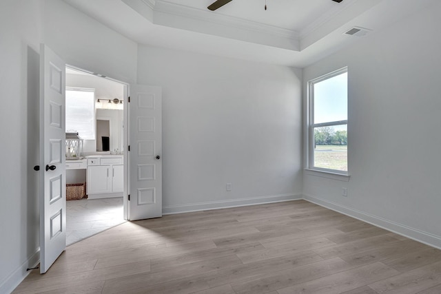 unfurnished bedroom featuring visible vents, ensuite bath, a tray ceiling, light wood-style floors, and crown molding