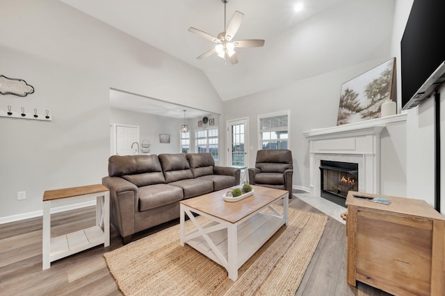 living room with baseboards, a ceiling fan, light wood-style flooring, a fireplace with flush hearth, and high vaulted ceiling