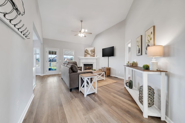 living room with ceiling fan, lofted ceiling, baseboards, light wood-style floors, and a lit fireplace