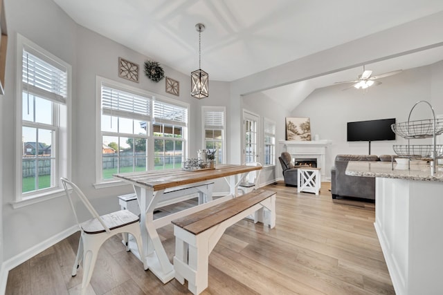 dining space with a warm lit fireplace, ceiling fan with notable chandelier, baseboards, vaulted ceiling, and light wood finished floors